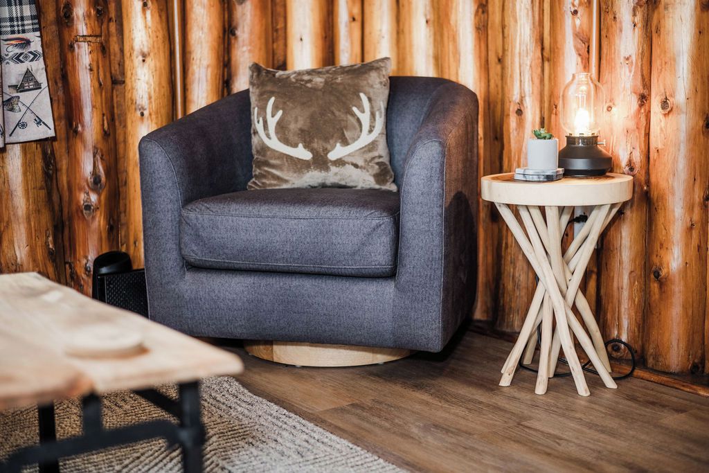Una habitación acogedora con un sillón gris, una pequeña mesita de madera con una lámpara y un panel de madera.