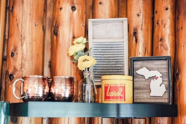 A wooden wall with a shelf holding a glass vase with yellow flowers, a metal kettle, and a framed map. - Vintage Lakefront Cabin-Fire Pit-Grill-Lake Views (BOYNE CITY)