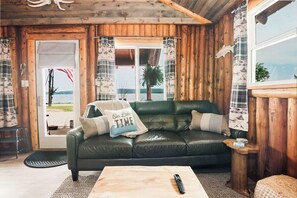 A cozy cabin interior with a leather sofa, a wooden coffee table, and a view of a lake through a door. - Vintage Lakefront Cabin-Fire Pit-Grill-Lake Views (BOYNE CITY)