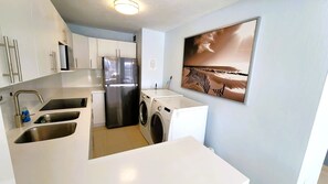 A modern kitchen with stainless steel appliances, a washing machine, and a large painting of a beach scene. - Ashford Avenue Beach Front Condo (San Juan)