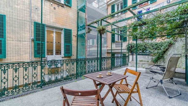 Un balcon avec une table et des chaises, une rambarde en métal et un immeuble aux volets verts.