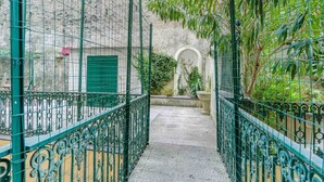 Un étroit sentier bordé de rampes de protection en métal ouvragé, menant à un bâtiment doté d’une porte verte et d’un arc décoratif.