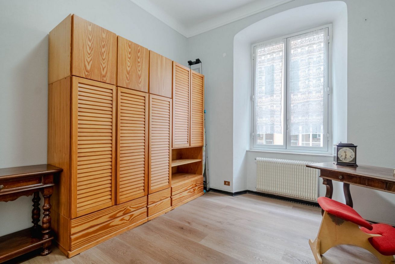 Een kamer met een houten garderobe, een radiator, een raam met zonwering, een bureau en een stoel.
