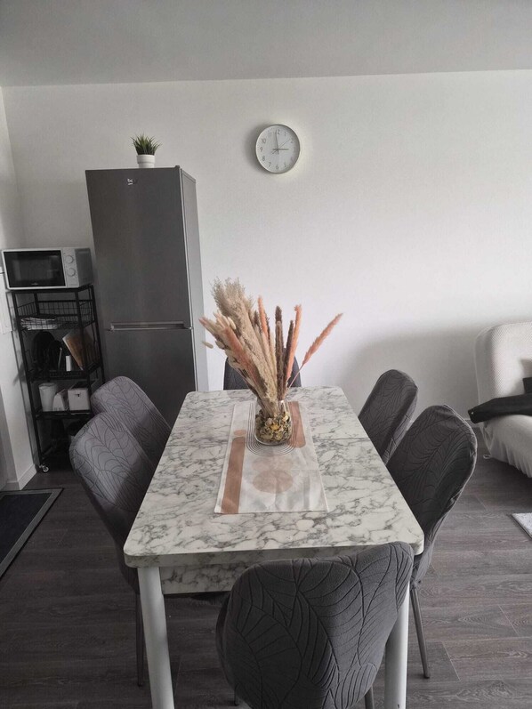 Un coin repas avec une table en marbre, des chaises, une horloge au mur, un réfrigérateur et une petite plante posée sur le dessus.