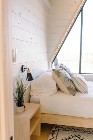 Un dormitorio con techo inclinado, cama con almohadas, lámpara de noche, mesita de madera con una planta y ventana con vistas.