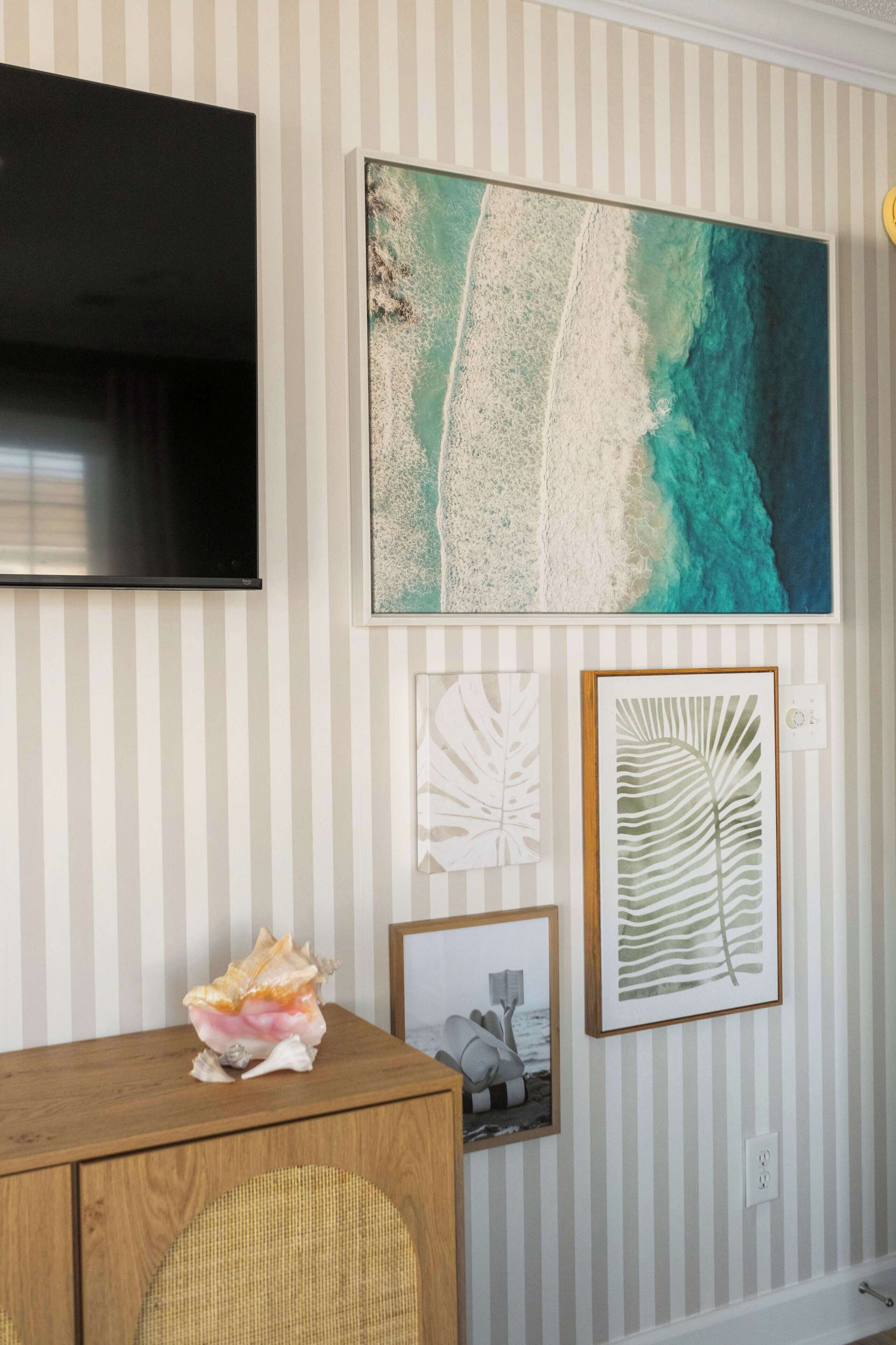 A room with a striped wall, a large framed ocean view painting, a wooden cabinet, and a television.