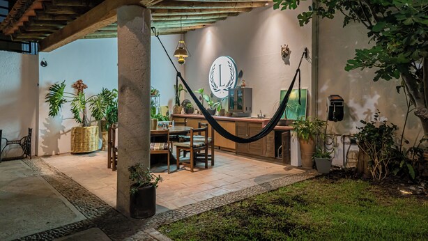 Een overdekt buitengebied met een hangmat, een eettafel met stoelen en potplanten.