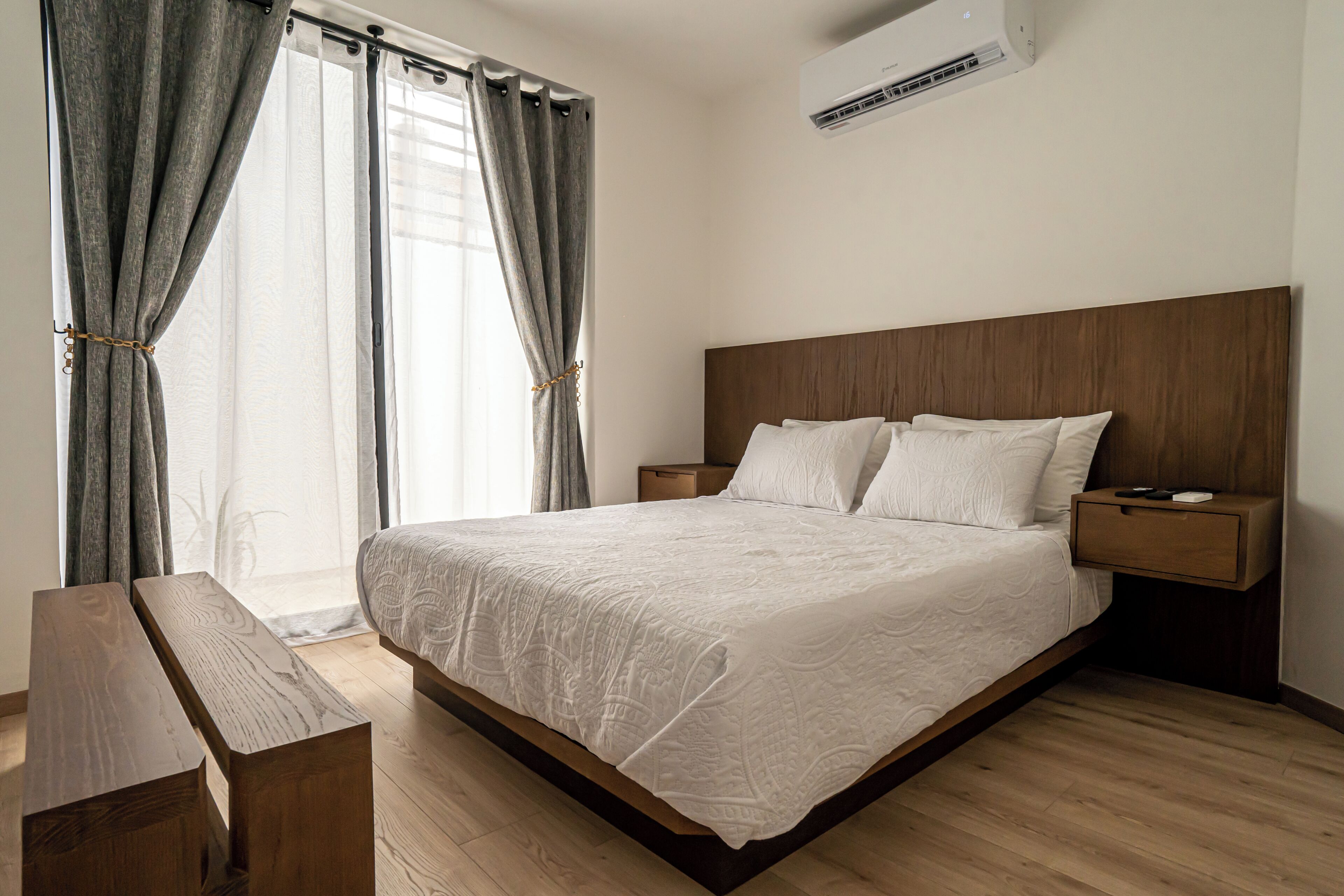 Un dormitorio con cabecera de madera, una cama con cubrecama blanco, un banco de madera, una mesita de noche con lámpara y un equipo de aire acondicionado.