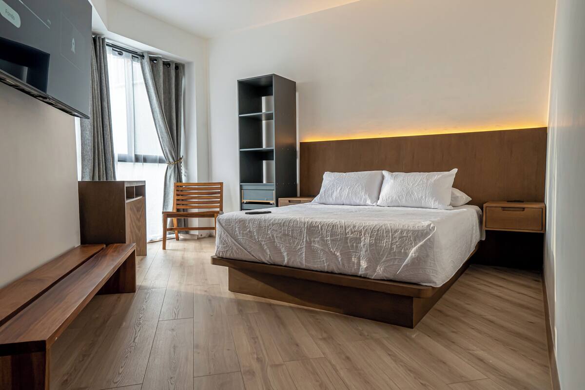 Una habitación de hotel moderna con una cama grande, suelo de madera, un banco, un televisor, una estantería y una mesita de noche.