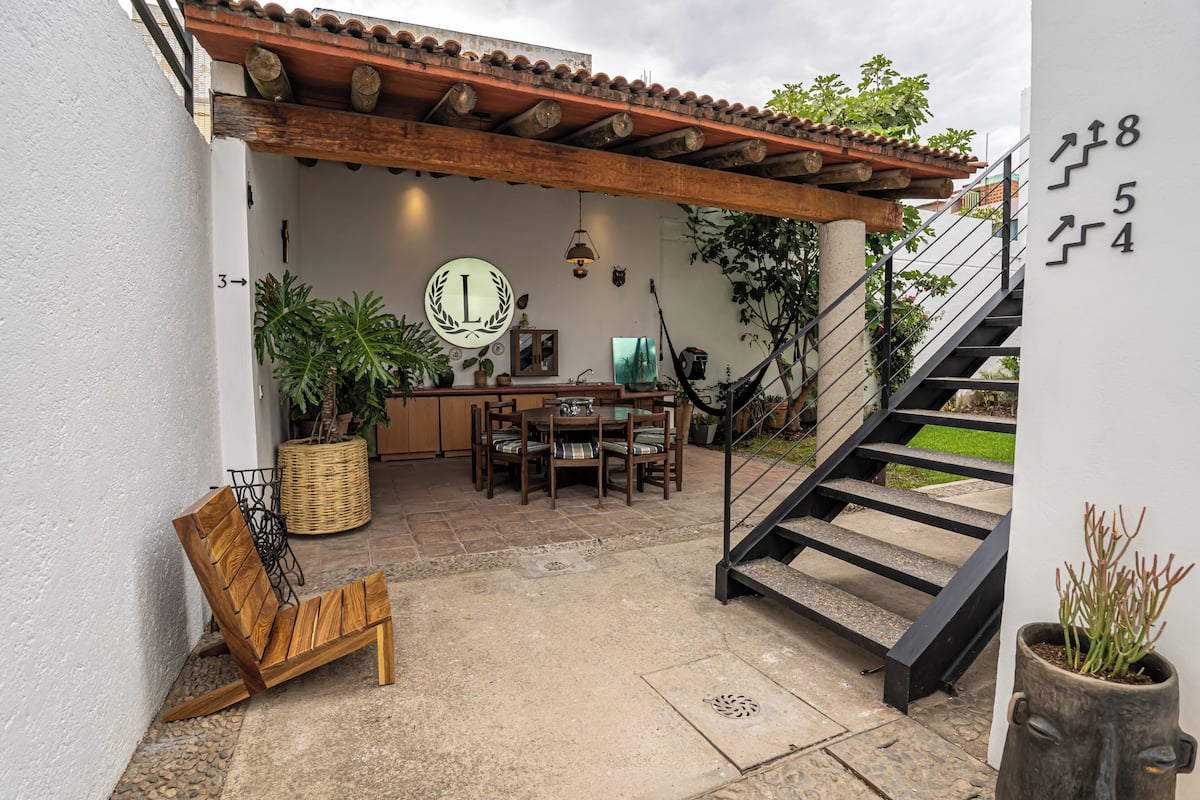 Un patio con una mesa y sillas de madera, una silla de madera, una cesta de mimbre y una escalera que conduce a un nivel superior.