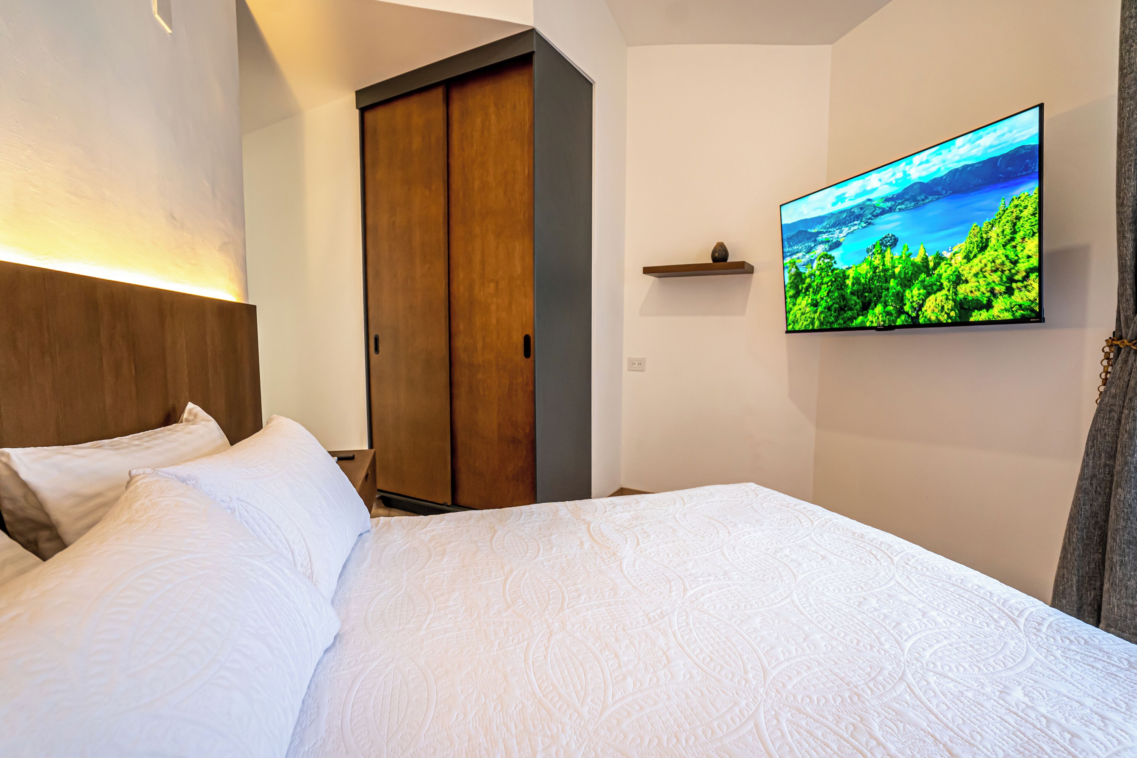 Una camera d'albergo con un letto, un armadio in legno, una TV a parete e uno scaffale con un oggetto decorativo.