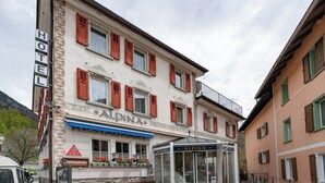 Ein Hotel mit der Aufschrift 'HOTEL ALPINA', einer Glastür, Sitzgelegenheiten im Freien und einem Bergpanorama im Hintergrund.