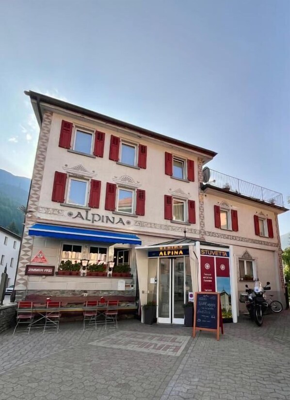 A two-story building with the name 'ALPINA' on the facade, a storefront with outdoor seating, and a signboard with 'STUETTA' visible. - Hotel Alpina (Zernez)