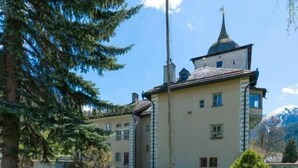 Ein zweistöckiges Gebäude mit einer symmetrischen Fassade, einem markanten Turm mit spitzer Dachform und einem gepflegten Rasen.