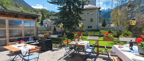 An outdoor patio with tables and chairs, a large tree, and a building with a mountain backdrop.