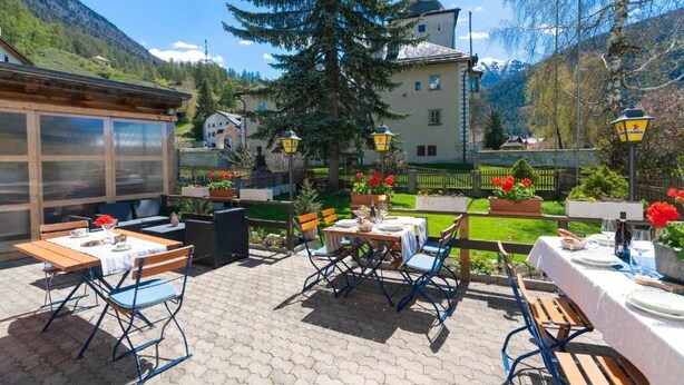 Eine Terrasse im Freien mit Tischen und Stühlen, im Hintergrund Berge und ein Gebäude mit einem Turm.