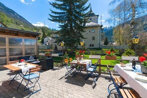 An outdoor patio with tables and chairs, a mountain backdrop, and a building with a tower. - Hotel Alpina (Zernez)