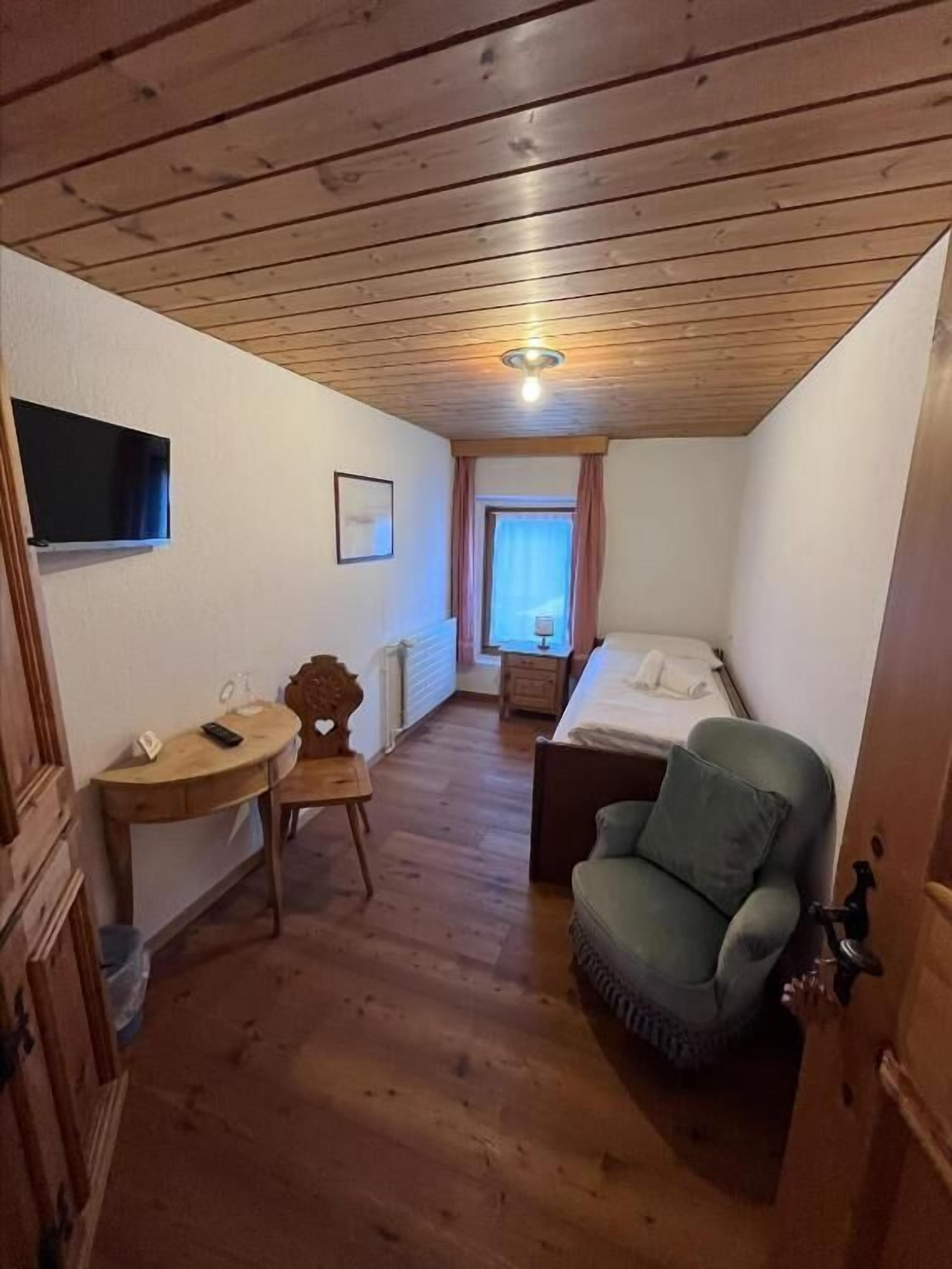 A small, cozy room with a wooden ceiling, a bed, a chair, a small table, and a TV.