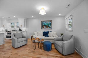 Ein modernes Wohnzimmer mit einem grauen Sofa, zwei Sesseln, einem Couchtisch und einem blauen Hocker. An der Wand hängt ein Gemälde, ein Fenster mit Jalousien und im Hintergrund eine Küche.