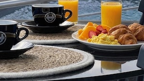 Colazione servita con una tazza di caffè, succo d'arancia, croissant e frutta su un tavolo vicino a una finestra con vista su una piscina e vegetazione.