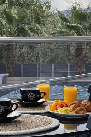 A breakfast setup with a coffee cup, orange juice, croissants, and fruit on a table by a window overlooking a pool and greenery. - Allure Villa, beachfront, featuring a gym, sauna, and jacuzzi (Porto Rafti)