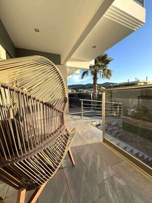 A modern outdoor balcony with a wicker chair, a glass railing, and a view of the cityscape. - Allure Villa, beachfront, featuring a gym, sauna, and jacuzzi (Porto Rafti)