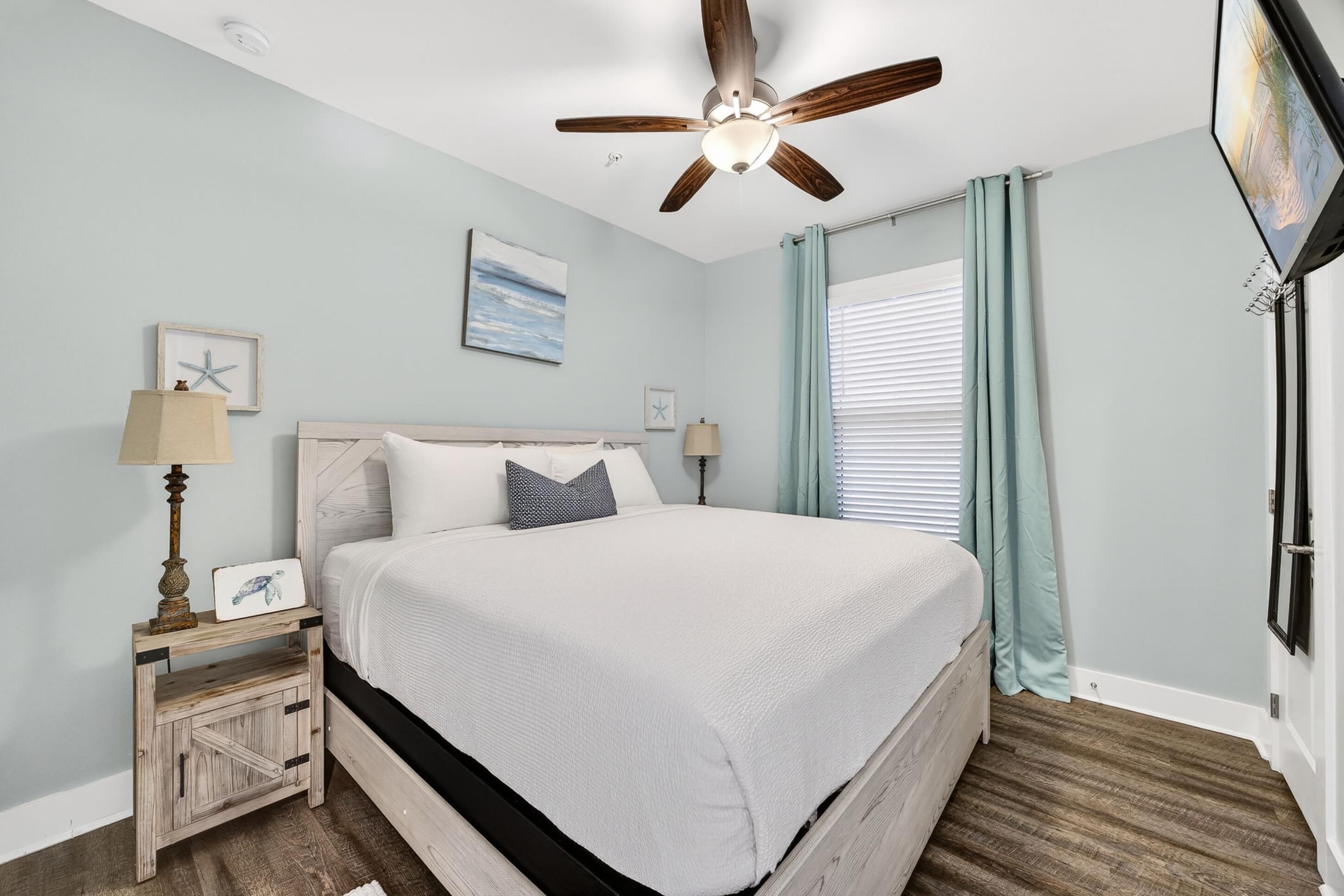 A bedroom with a bed, a ceiling fan, a nightstand, a lamp, a window with curtains, and a wall-mounted TV.