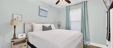A bedroom with a bed, a ceiling fan, a nightstand, a lamp, a window with curtains, and a wall-mounted TV.