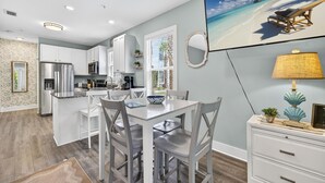 A modern kitchen with white cabinetry, a dining area with a white table and chairs, and a beach view painting on the wall. - 30A - Hidden Lakes Condo Rentals (Santa Rosa Beach)