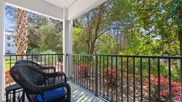 A covered balcony with wicker furniture, a railing, and a view of trees and shrubs.