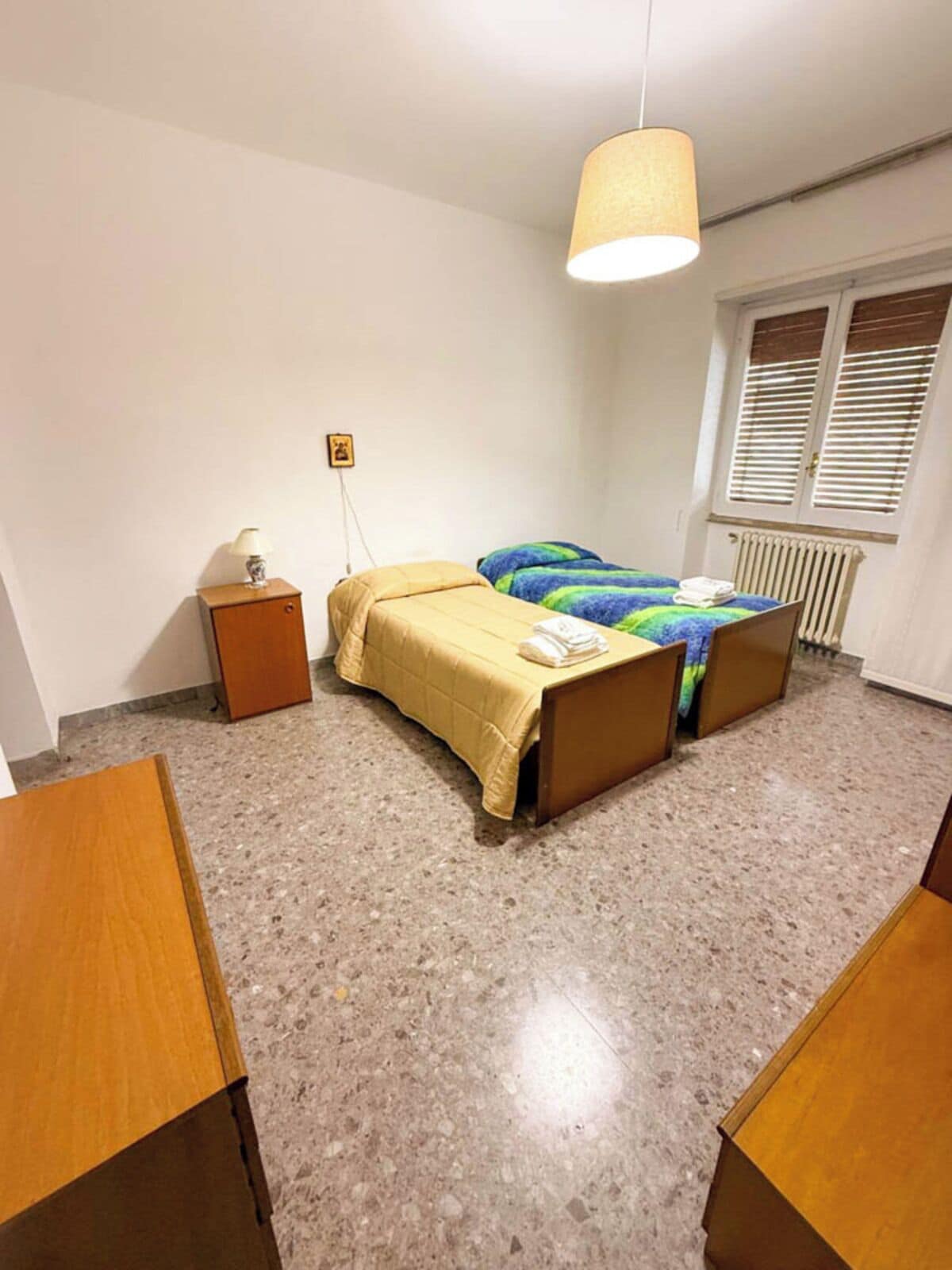 A bedroom with a bed, a bedside table, a lamp, a radiator, and a window with blinds.