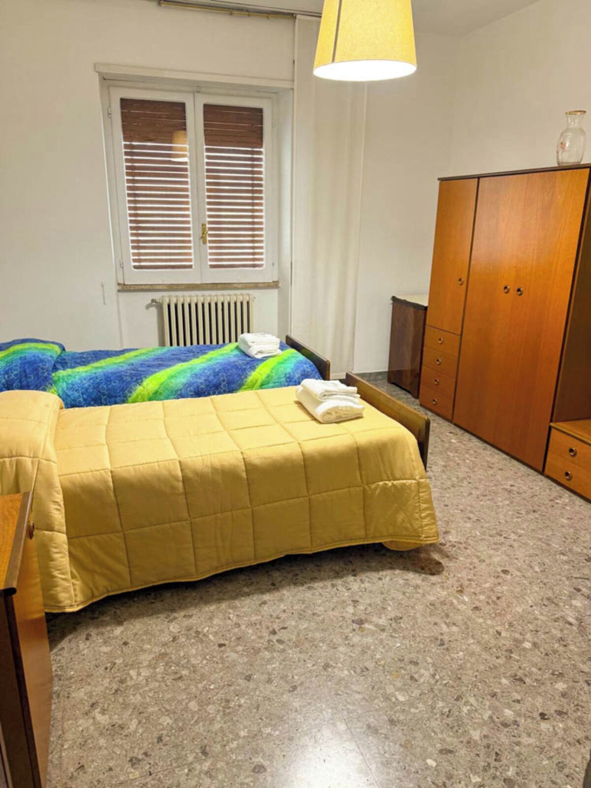 A room with a bed, a wardrobe, a radiator, and a window with blinds.