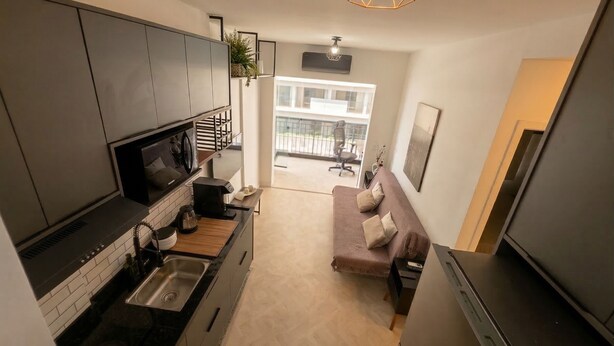 A compact kitchen with a built-in oven, a coffee machine, and a sink. There is a sofa with cushions in the living area, and a balcony with a railing is visible through the window. - Cozy studio near Paulista Avenue (São Paulo)