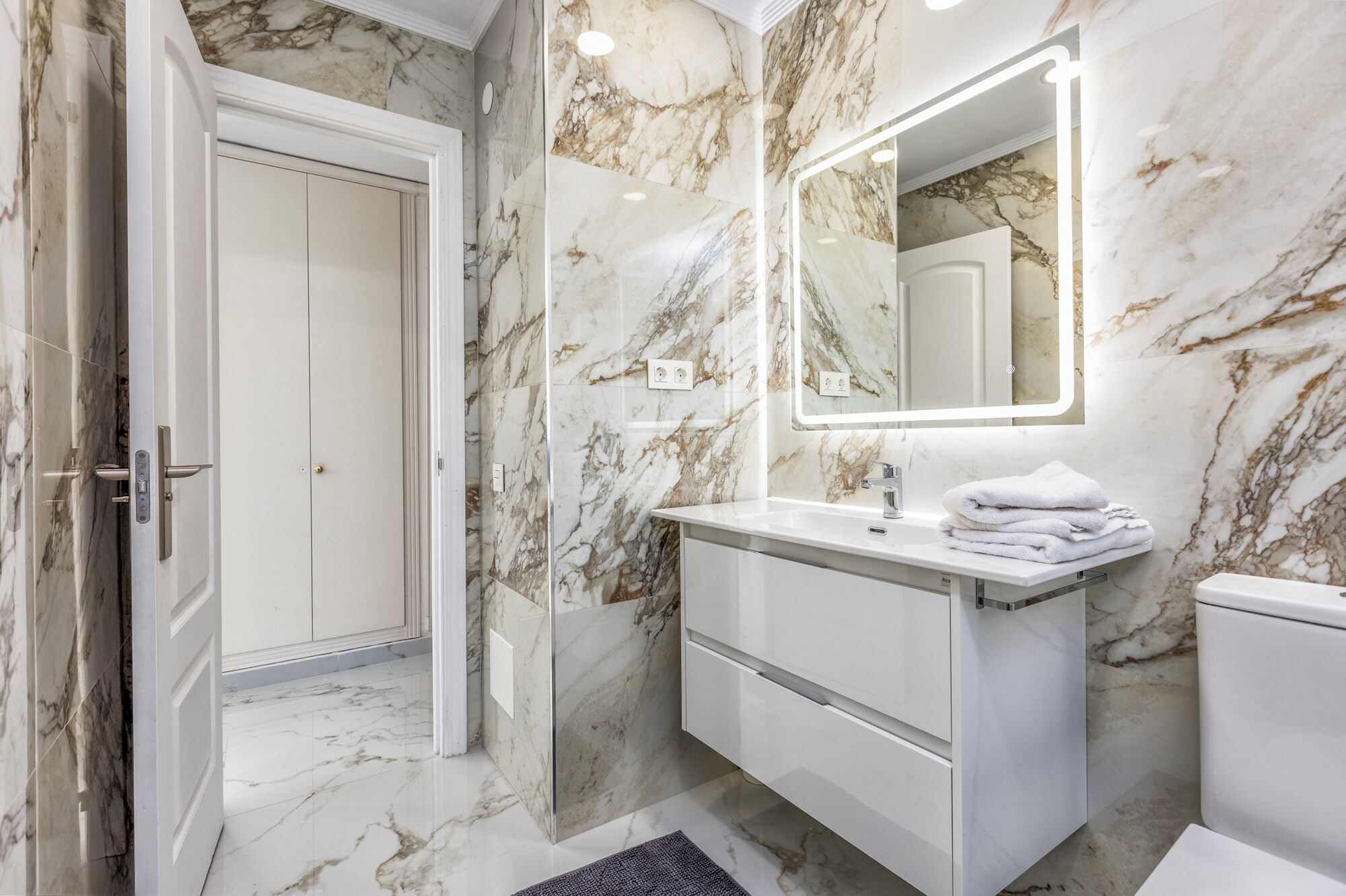 A bathroom with marble walls, a white vanity, a mirror with lights, and a door.