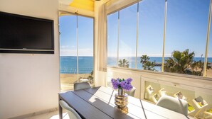 Ein Zimmer mit großem Fenster mit Meerblick, einem an der Wand montierten Flachbildfernseher, einem Esstisch mit Stühlen und einer Vase mit Blumen.