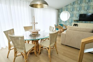 Ein Wohnzimmer mit einem Esstisch aus Glas, umgeben von Korbstühlen, einem beigen Sofa und einem an der Wand befestigten Fernseher.