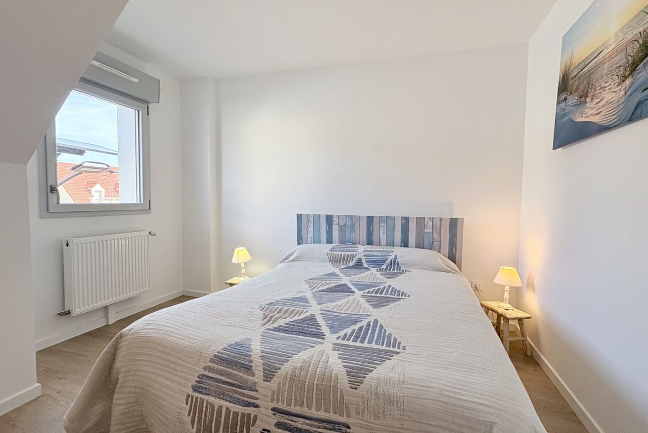 A bedroom with a bed, two bedside lamps, a window, a radiator, and a painting on the wall.