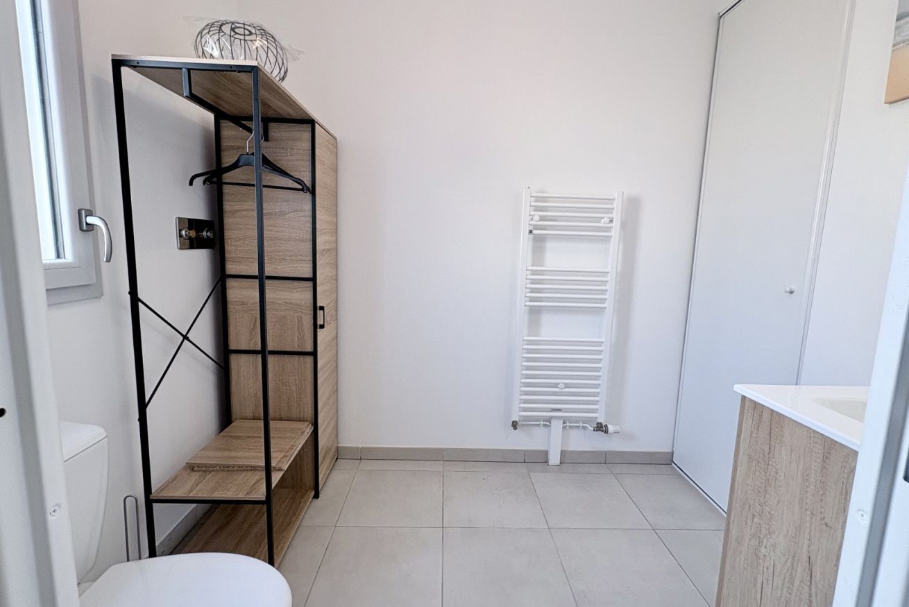 Un baño con inodoro blanco, una estantería de madera con ropa colgada y un radiador blanco.