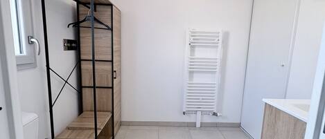 Un baño con inodoro blanco, una estantería de madera con ropa colgada y un radiador blanco.