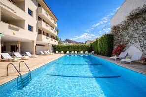 Piscina com espreguiçadeiras, um edifício com varandas e um céu claro.