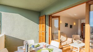 A balcony with a table set for two, a white wicker chair, a sofa, and a sliding glass door leading to an indoor living area. - Apartment 2C - Penthouse Bressol (Port de Pollença)