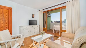A living room with a TV, white furniture, a table with a vase, and a sliding glass door to an outdoor area. - Apartment 2C - Penthouse Bressol (Port de Pollença)