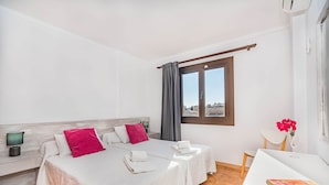 A hotel room with two beds, a desk, a chair, a window with curtains, and a view of buildings outside. - Apartment 2C - Penthouse Bressol (Port de Pollença)