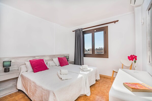 A hotel room with two beds, a desk, a chair, a window with curtains, and a view of buildings outside. - Apartment 2C - Penthouse Bressol (Port de Pollença)