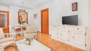 A living room with a TV, white furniture, a dining table, and a wooden door. - Apartment 2C - Penthouse Bressol (Port de Pollença)