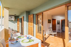 A balcony with a table set for dining, wooden shutters, and a sliding glass door leading to an interior room with a chair and a painting. - Apartment 2B - Penthouse Bressol (Port de Pollença)