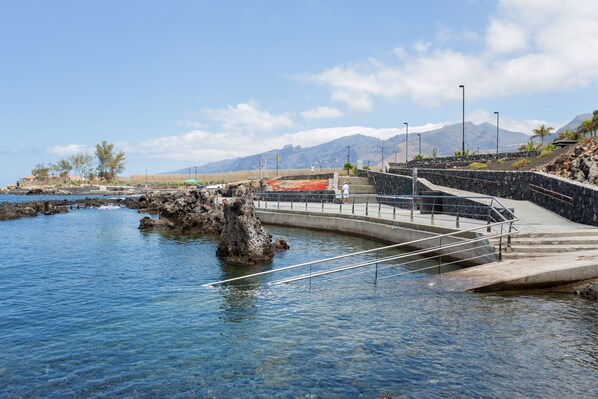 Une promenade en bord de mer agrémentée d’une balustrade en pierre, avec une eau d’un bleu limpide et une chaîne de montagnes en arrière-plan.