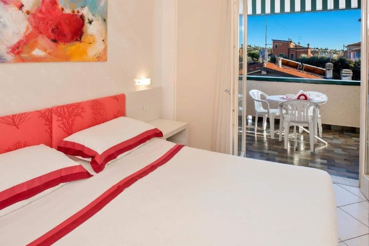 A hotel room with a bed, a painting, a balcony with a table and chairs, and a view of buildings.