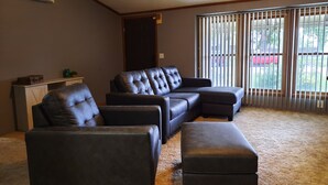 Ein Wohnzimmer mit Ecksofa, zwei Sessel, einem Couchtisch, einer kleinen Kommode und einer Glastür zum Balkon.