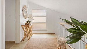 Un pasillo con suelo de madera, pared blanca, una ventana, una planta y una lámpara en el techo.